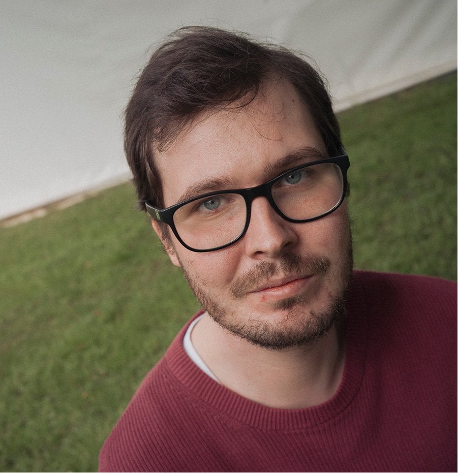 Mężczyzna z brązowymi włosami i czarnymi okularami na tle zielonej trawy.